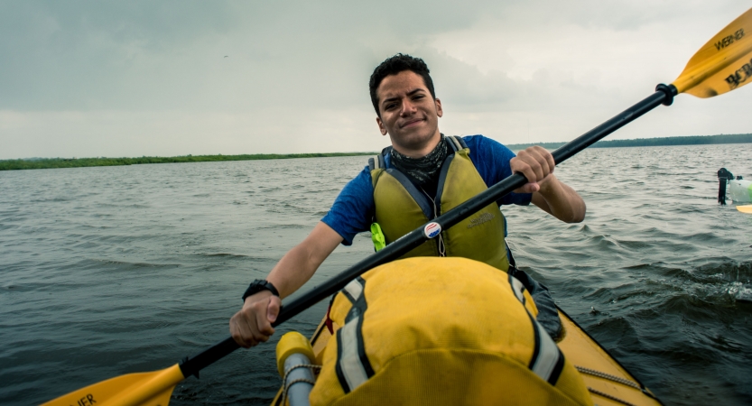 A young person smiles at the camera while paddling on a large body of water. 